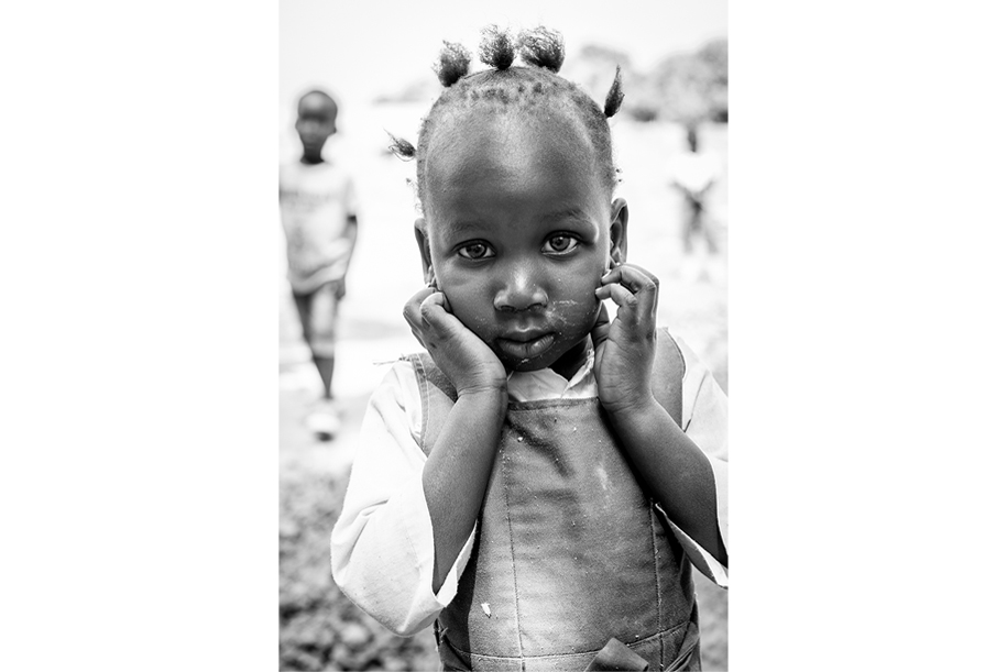 Black and white portrait of a girl from Gambia. Looking shy into the camera, with both hands on her cheeks. Ellis Photography