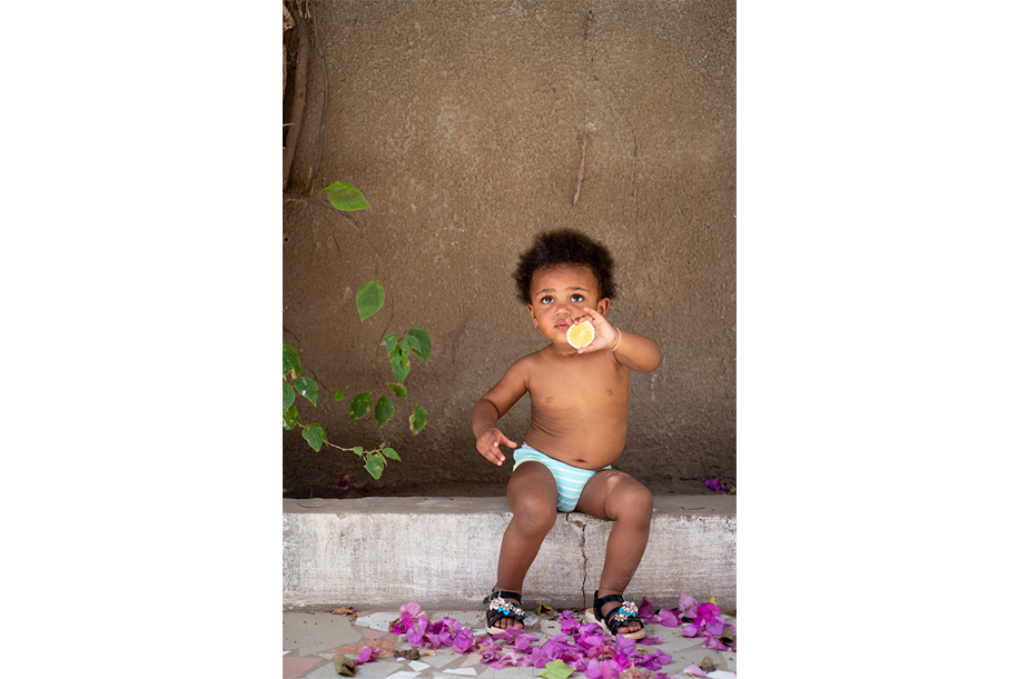 Cute little girl sitting with an orange in her hand. Pink flowers on the floor, plain wall and some green leaves. Kids photography by Ellis